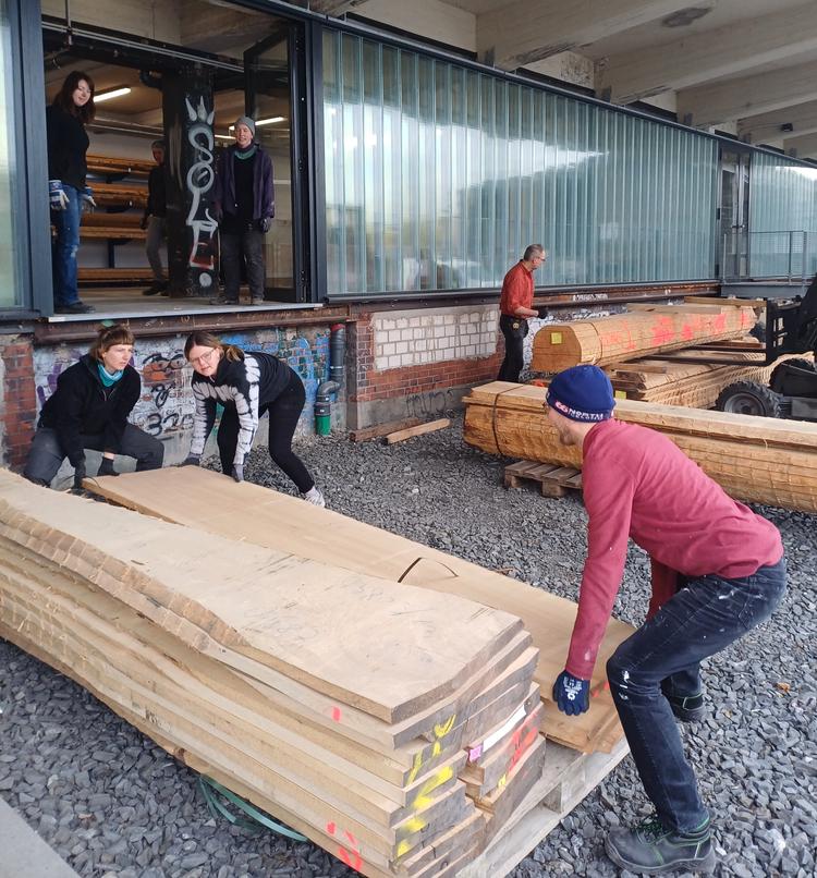 Eine große Holzlieferung ist in der B-Side angekommen und wird von vielen Menschen in die Holzwerkstatt getragen.