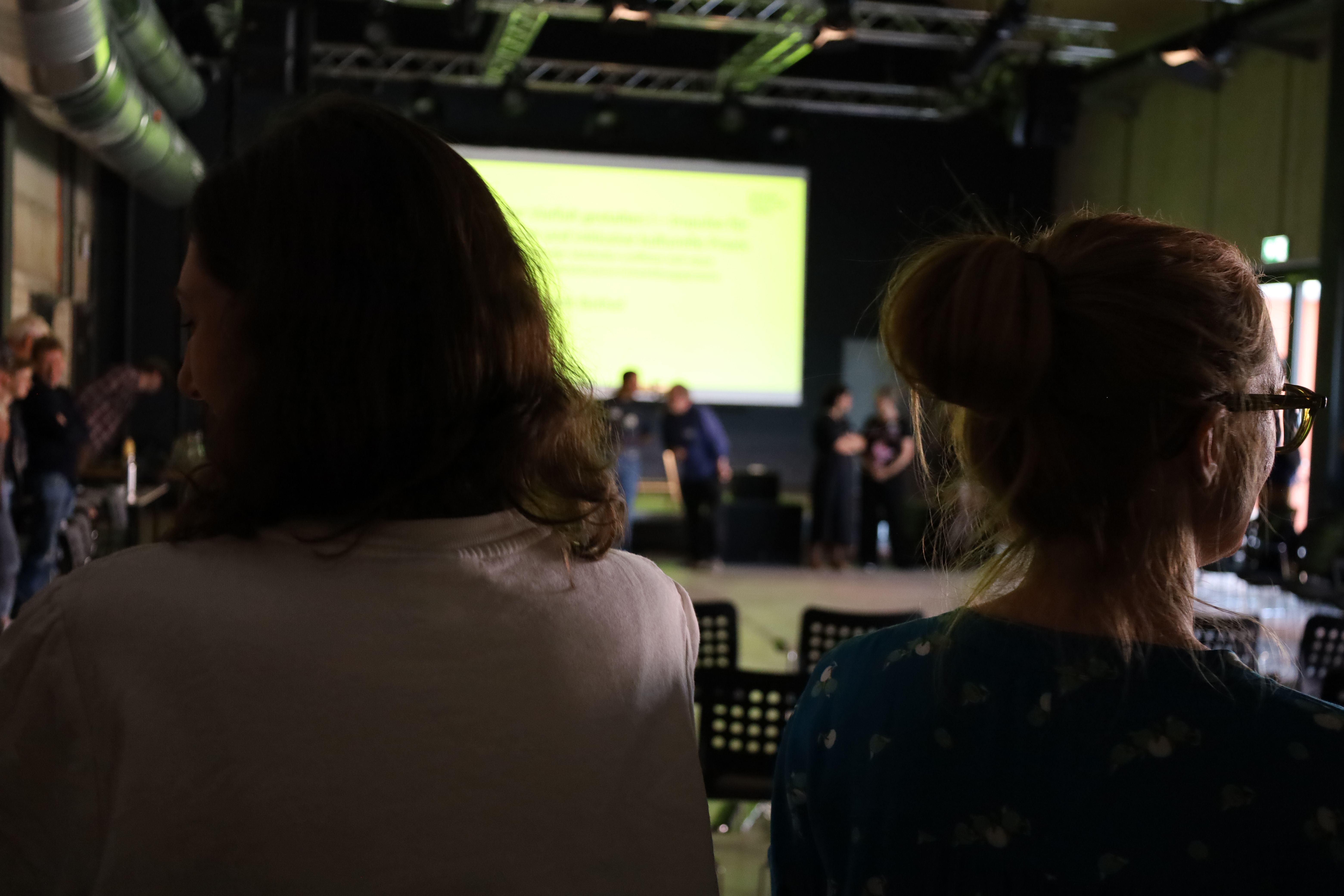 Ein Foto aus dem Veranstaltungsraum. Menschen bilden einen Kreis und unterhalten sich. Im Hintergrund ist ein Leinwand zu sehen. Weder Menschen noch der Inhalt der Leinwand ist erkennbar.