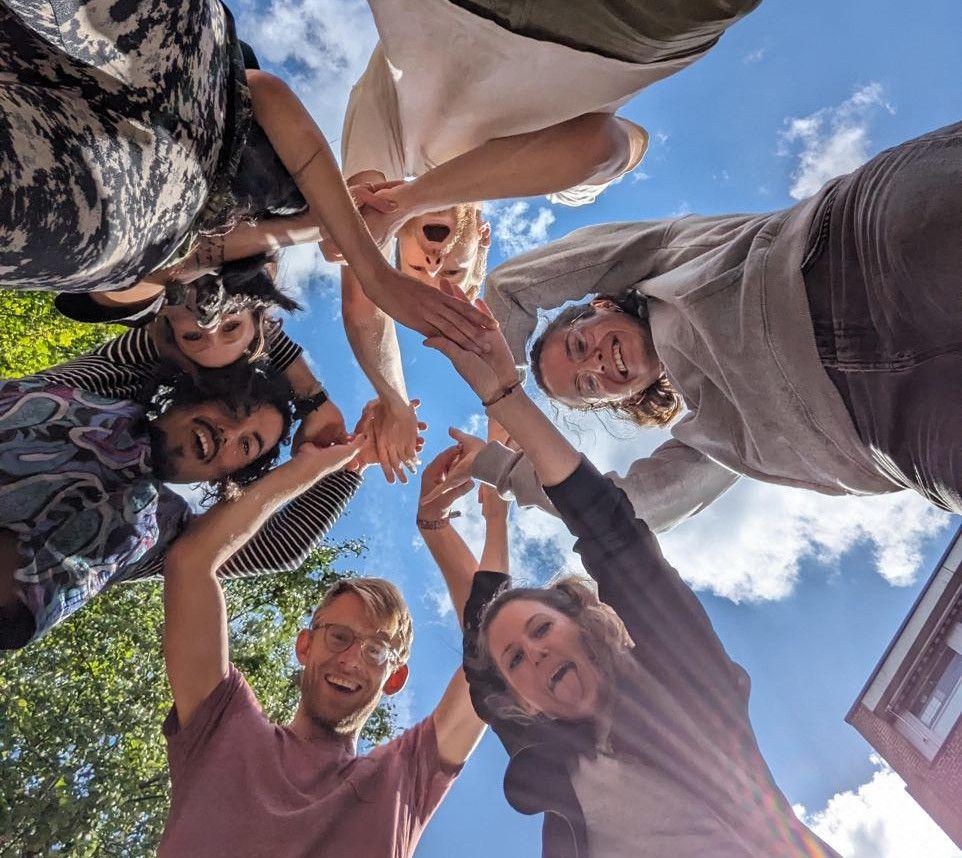 Die Kamera fotografiert von unten in den Himmel. Über der Kamera stehen Übungsleiter*innen des Freiraum-Kollektivs herum und lächeln mit erhobenen Händen in die Kamera.