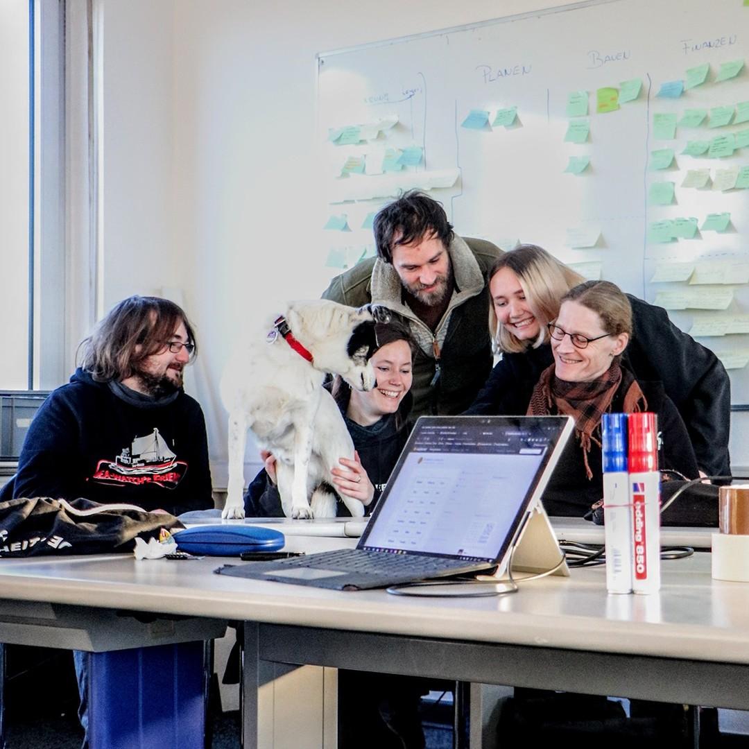 Team-Foto des Öffi-Teams. Fünf Menschen sitzen an einem Tisch und schauen auf einen hinter einem Laptop verdeckten Gegenstand.